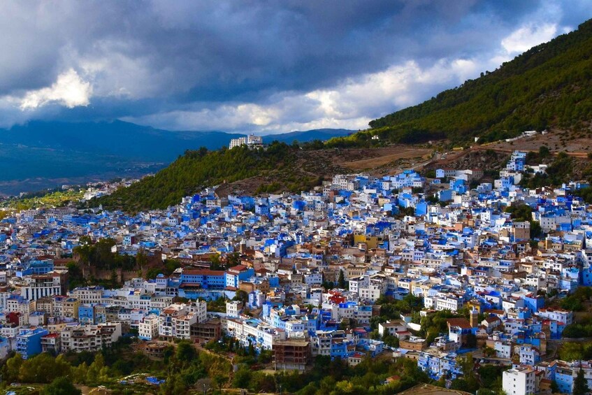 A densely packed town with buildings predominantly painted in blue and white, set against a backdrop of mountains and cloudy skies. 5