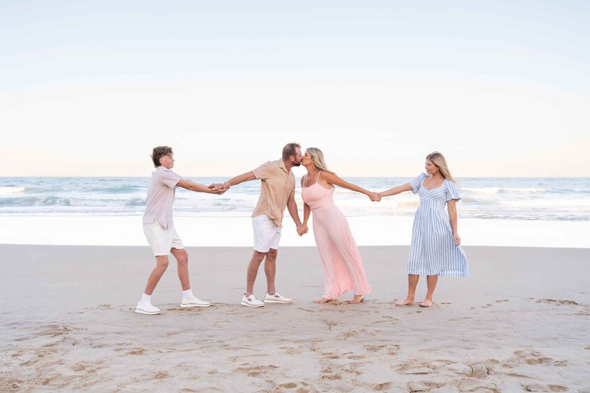 A family of four holding hands on a sandy beach with the ocean in the background. 1