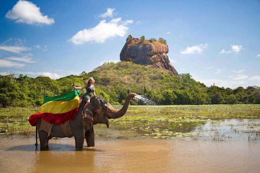 An elephant with a rider in a natural setting, a large rock formation in the background, and a body of water. 1