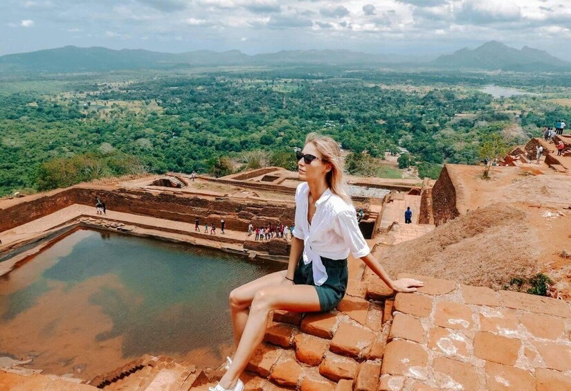 A person sitting on a stone ledge overlooking a historical site with a large water body and distant mountains. 2