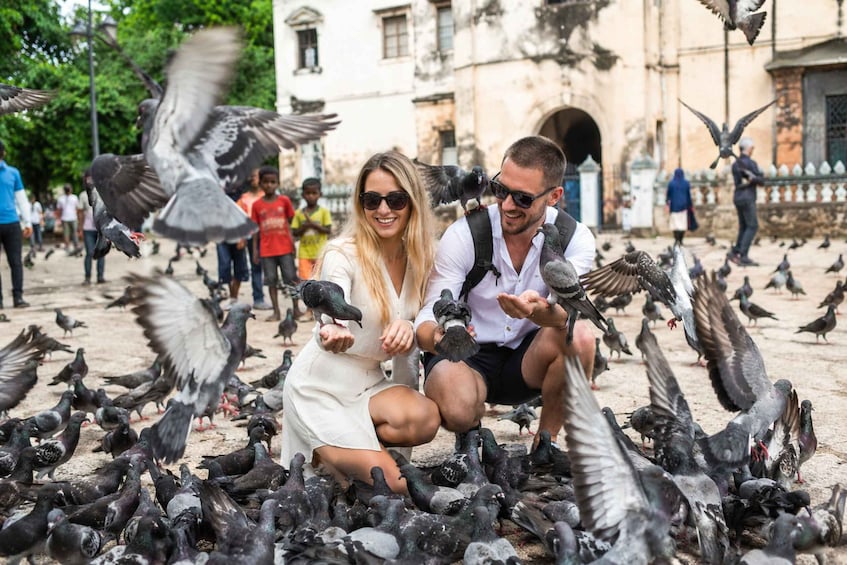 A couple feeding pigeons in a public square with historic buildings in the background. 2