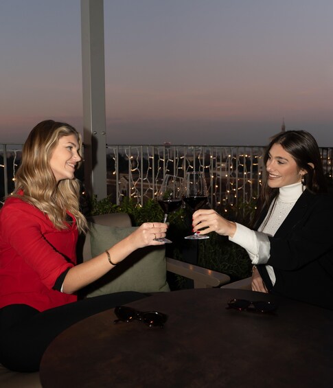 Two people toasting with wine glasses at an outdoor table with a cityscape in the background. 1