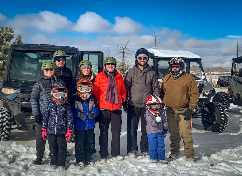A group of people in winter clothing and helmets standing in snow with off-road vehicles in the background. 3