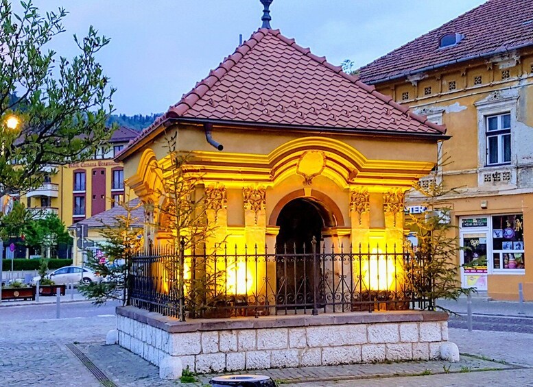 A small, ornate building with a tiled roof and a stone base, surrounded by a wrought iron fence and trees. 5