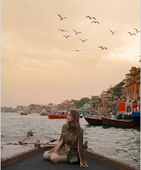 A person sitting on a boat with birds flying overhead, a river, and buildings in the background. 4