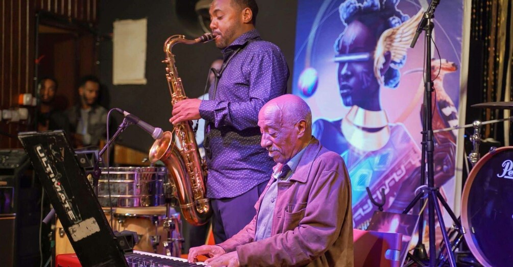 A saxophonist and a keyboard player performing on stage with musical instruments and a drum set. 1