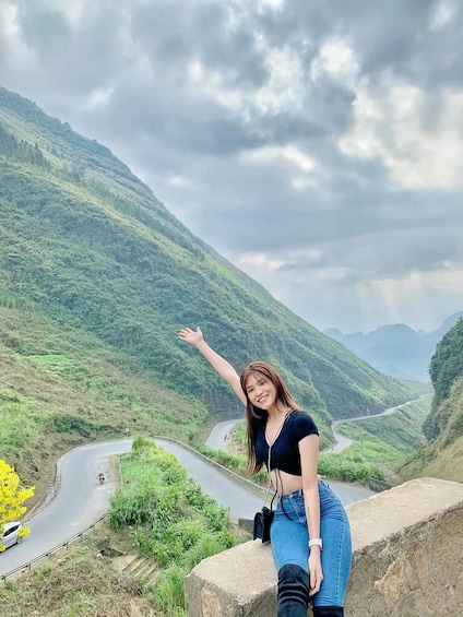 A woman standing on a stone ledge overlooking a winding road through a mountainous landscape. 4