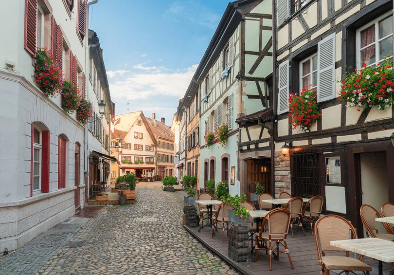 A cobblestone street lined with traditional half-timbered buildings, outdoor seating, and hanging flower baskets. 1