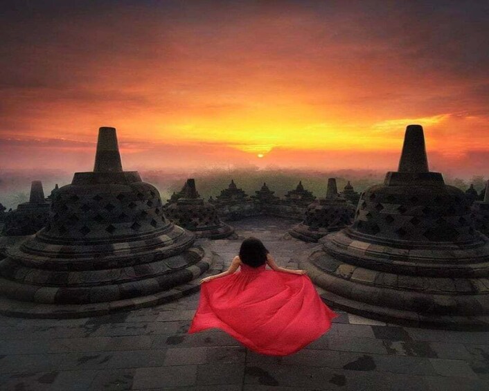 A person in a red dress standing on a stone platform with a sunset in the background. 1