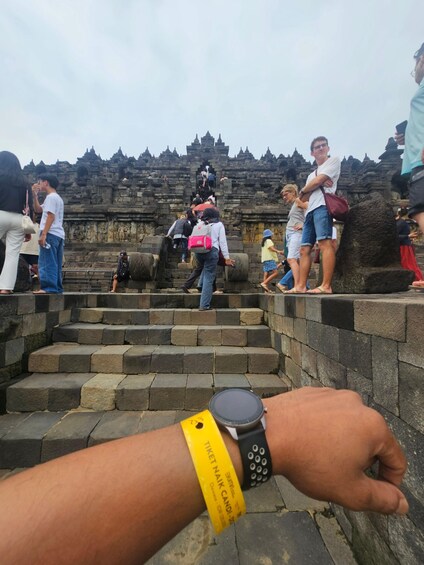 A person wearing a wristband at a temple entrance with stone steps and visitors. 5