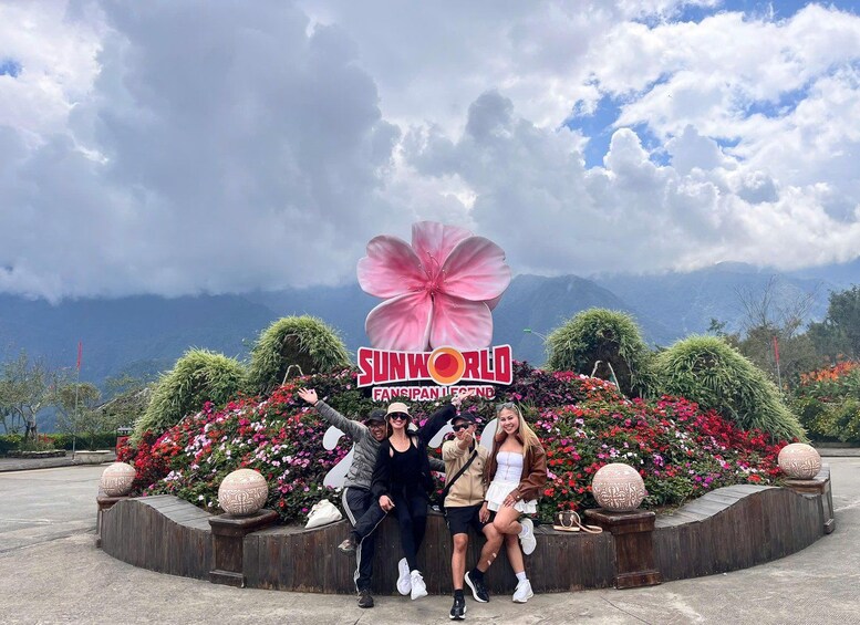 A group of people posing in front of a 'SUNWORLD FANSIPAN LEGEND' sign with a large pink flower decoration, surrounded by colorful flowers and plants. 3