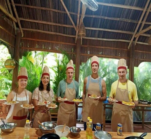 A group of people in a rustic kitchen setting, wearing chef hats and aprons, holding plates of food. 3