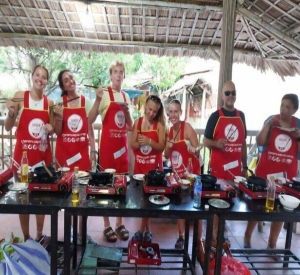 A group of people wearing red aprons standing around a table with cooking equipment and ingredients. 1