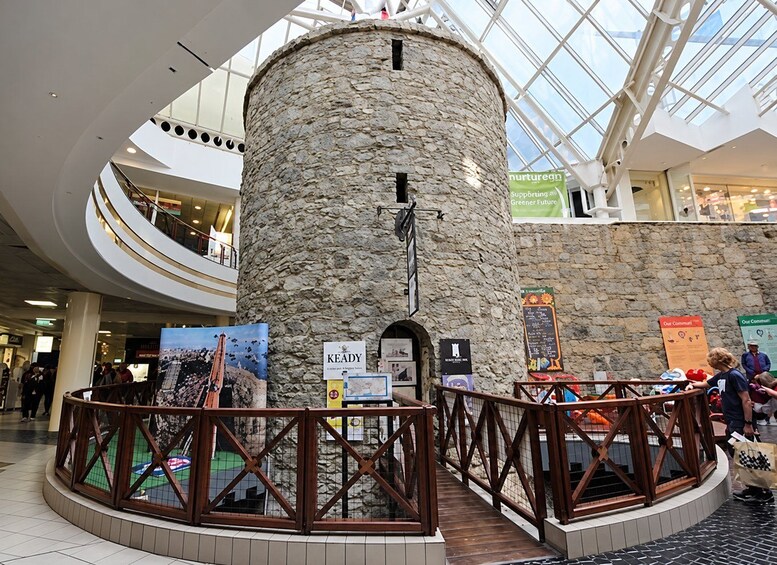 A stone tower with a cross, surrounded by a wooden fence, inside a modern building with glass ceilings and multiple levels. 2