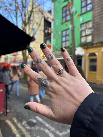 A hand with green-painted nails and multiple tattoos, including a turtle and a flower, against a blurred urban street background. 3