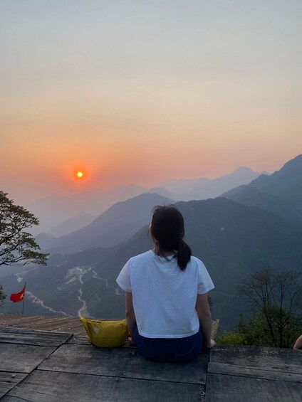 A person sitting on a wooden platform overlooking a valley with mountains and a river, during sunset. 3