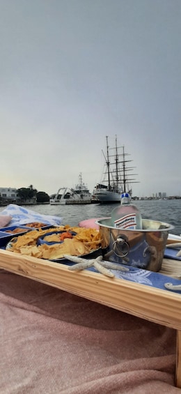 A wooden table with snacks and a bottle of liquor by a waterfront with a tall ship in the background. 5