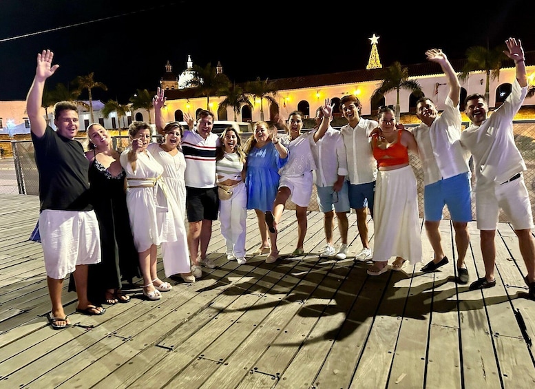 A group of people on a wooden boardwalk at night, with a building and palm trees in the background. 2