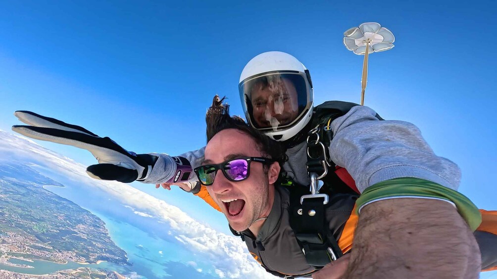 A person skydiving with a parachute, another person in a helmet, and a landscape with clouds and water below. 1