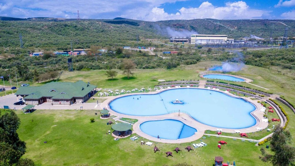 A large outdoor swimming pool complex surrounded by green lawns and a few buildings. 1