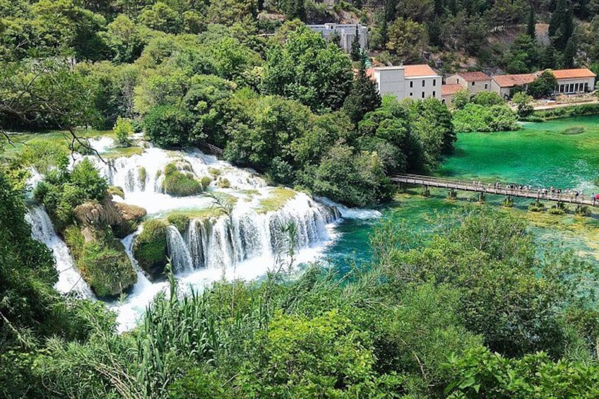 A waterfall cascading through a lush green landscape with a bridge and buildings in the background. 2