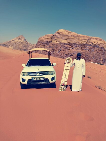 A white car with a license plate in a desert with red sand and a large rock formation in the background. 1