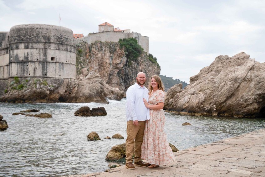A couple standing by a rocky waterfront with a historic fort in the background. 2