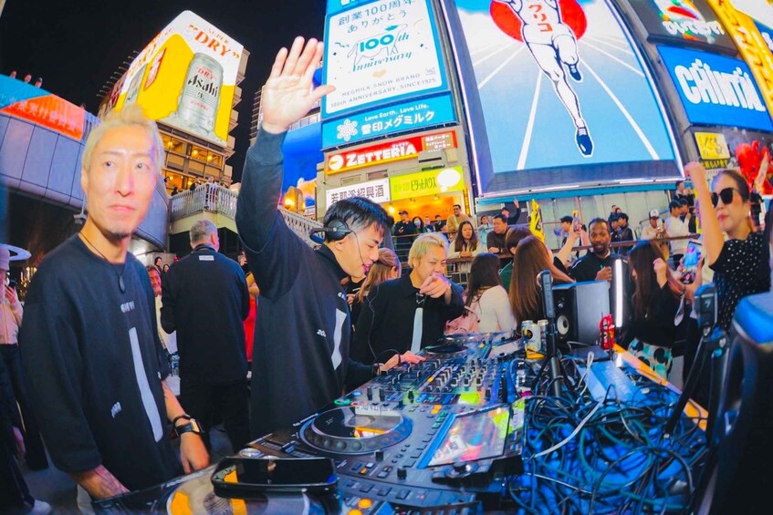 A crowded urban night scene with illuminated billboards and a DJ booth. 2