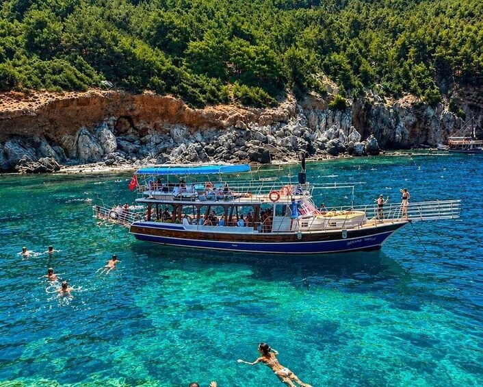 A boat with people swimming in clear blue water, surrounded by rocky cliffs and greenery. 1