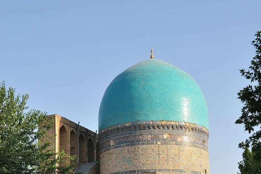 A large turquoise dome with intricate patterns and Arabic calligraphy, surrounded by trees and a brick structure. 3