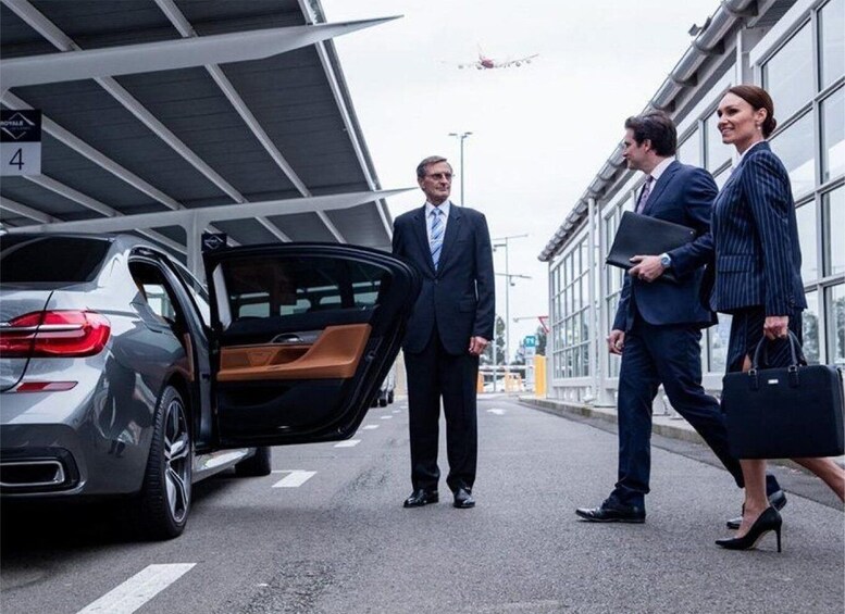 A man in a suit walking towards a car with its door open, a woman in a suit walking beside him, and another man holding a briefcase. 1