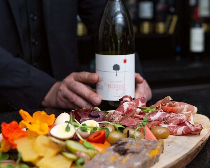 A person holding a bottle of rosso wine with a charcuterie board in front. 2