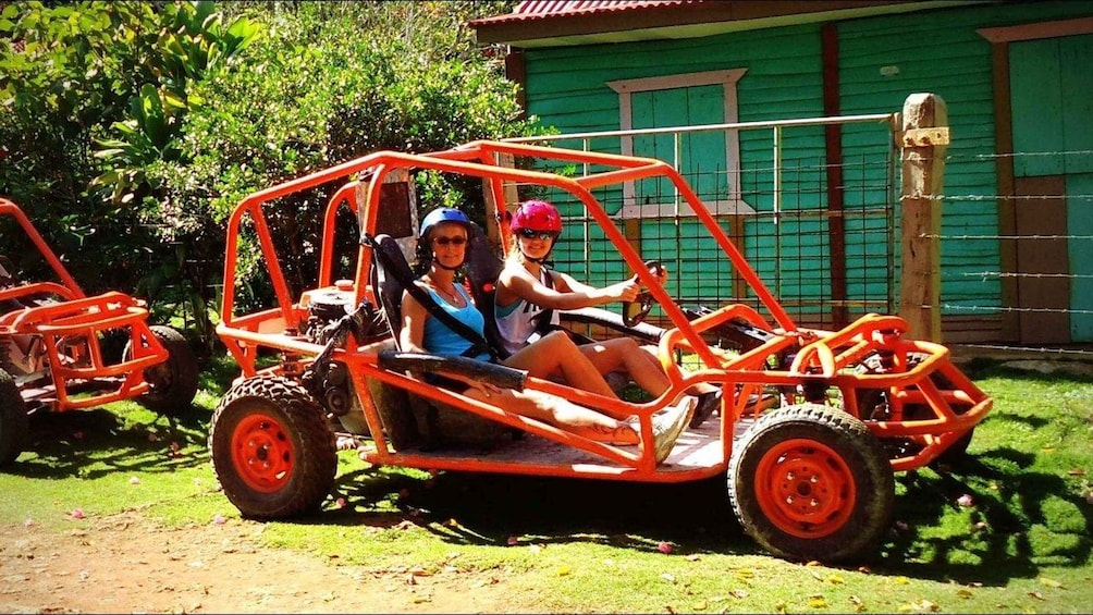 Two people in a red off-road vehicle on a dirt path with green foliage and a green building in the background. 5