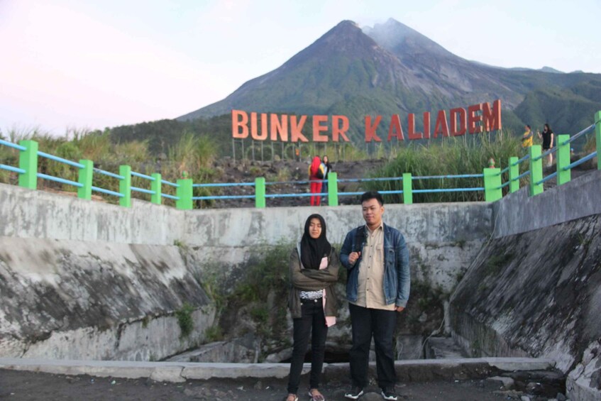 A couple standing in front of a sign that reads "BUNKER KALIADEN" with a mountain in the background. 2