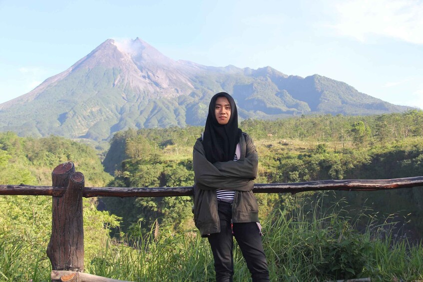 A person standing with arms crossed in front of a wooden fence, overlooking a lush green landscape with a prominent mountain in the background. 3