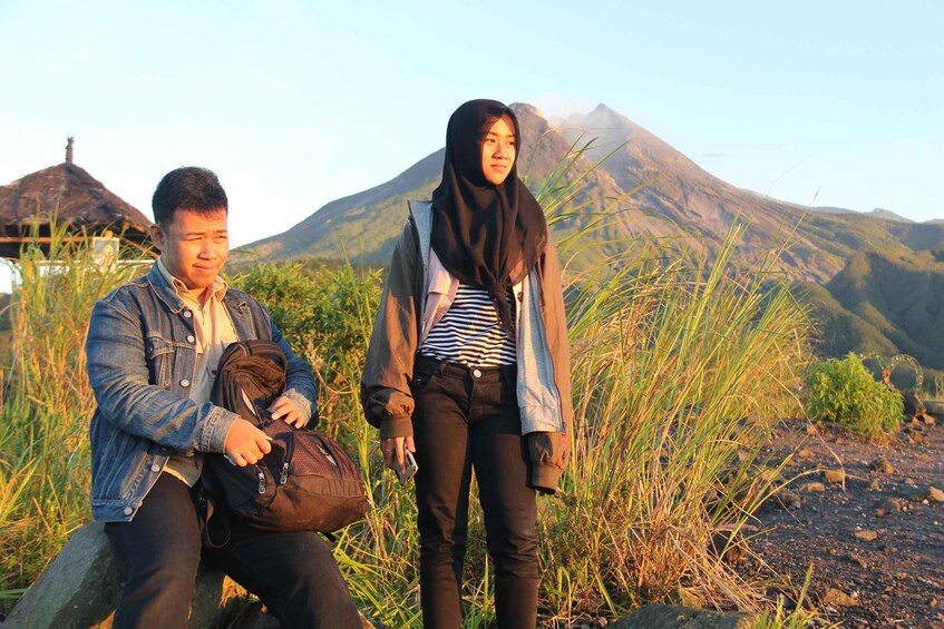 Two people sitting and standing in a grassy area with a mountain in the background. 4