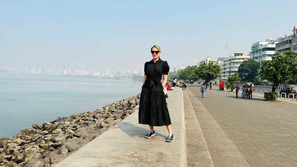 A woman in a black dress standing on a concrete path by a rocky shoreline with a city skyline in the background. 5