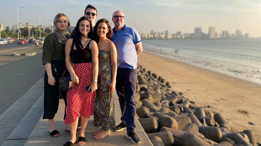 A group of people standing on a concrete walkway by a rocky beach with a city skyline in the background. 1