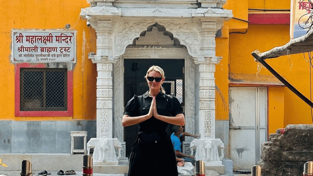 A person in black prayer pose in front of a temple with a sign in a local language. 2