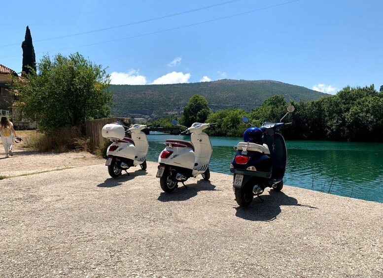 Two scooters parked on a gravel path by a lake with a hill in the background. 4