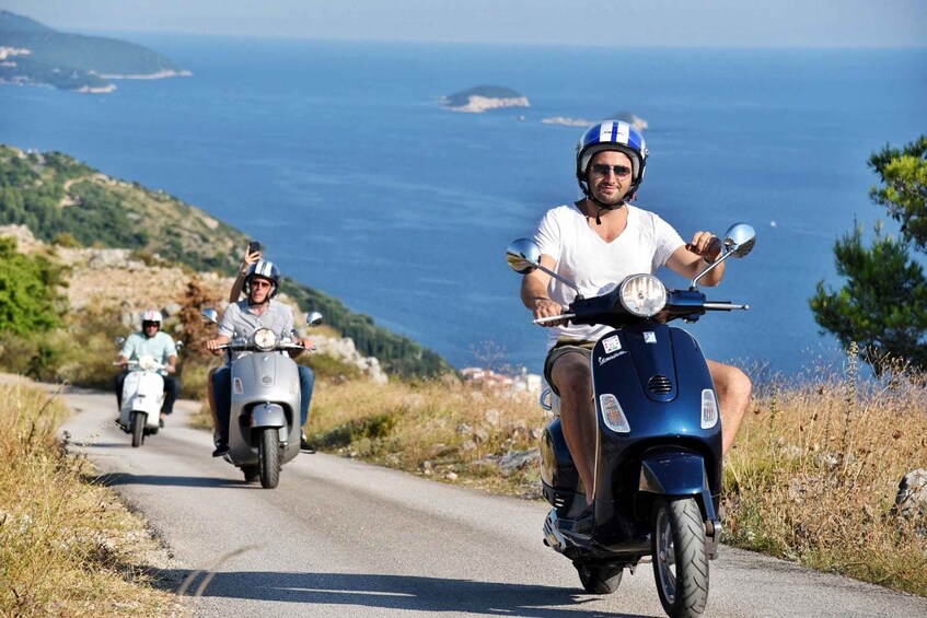 Three people riding scooters on a coastal road with a view of the sea and hills in the background. 5