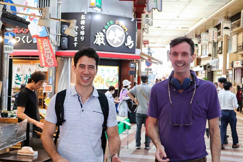 Two men standing in a busy market with various shops and signs in the background. 1
