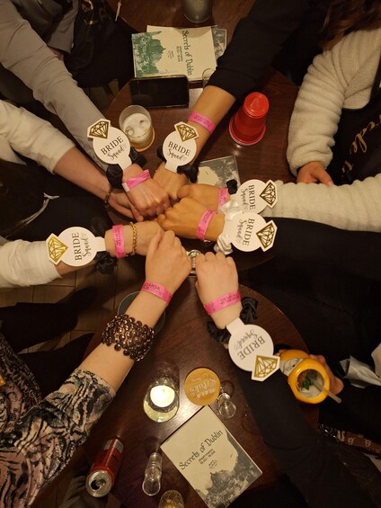A group of people sitting around a table with their hands joined in the center, wearing bracelets with 'BRIDE SQUAD' tags. 4