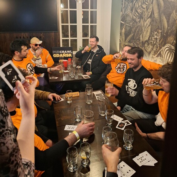 A group of people seated around a table with glasses of beer, playing cards, and a sign that reads 'PUBCRAWL GDansk'. 2