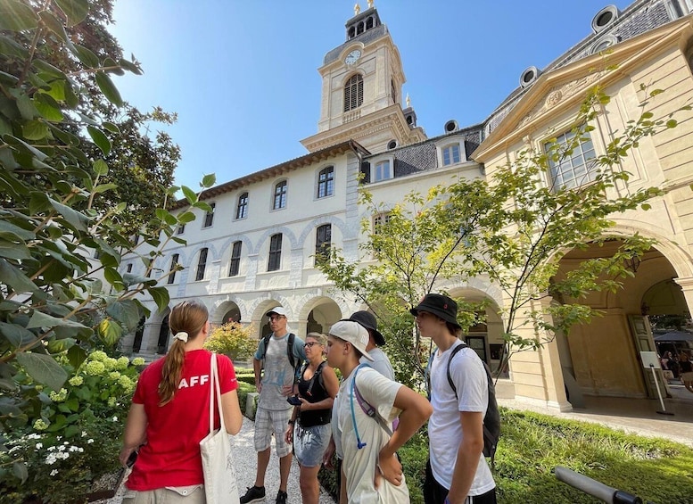 A group of people standing in a courtyard with a historic building featuring arches and a clock tower in the background. 1