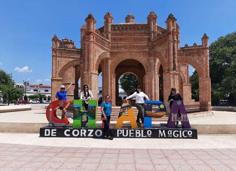A group of people posing in front of a historic brick archway with the text 'De Corzo Pueblo Magico' below. 3