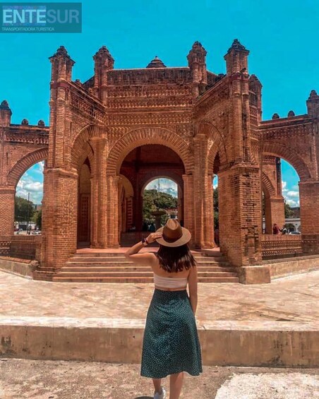 A woman in a hat and dress standing in front of a large, ornate brick archway. 2
