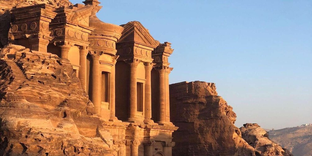 A large sandstone structure with columns and intricate carvings, set against a rocky cliff. 1