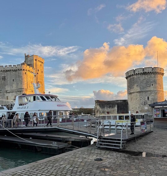 A historic castle with a round tower, a white boat named 'ORAZUR 3' docked at a pier, and a cobblestone waterfront. 2