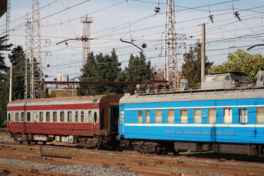 A train with a red and blue carriages on tracks, surrounded by electrical towers and trees. 3